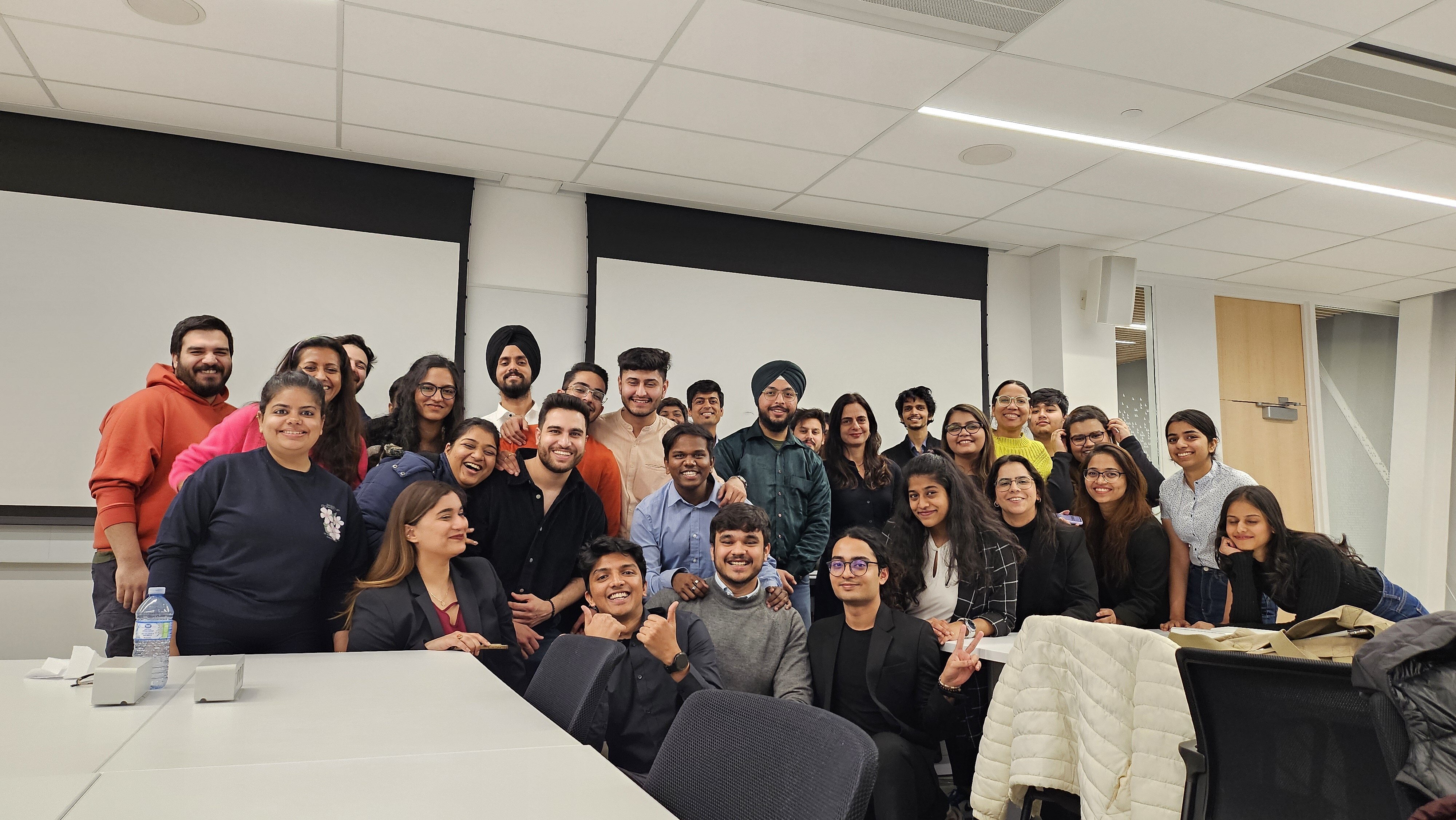 A group of students from York University, School of Continuing Studies, posing together in a classroom. The diverse group is smiling and showing camaraderie, with some students giving thumbs-up and peace signs, creating a cheerful and inclusive atmosphere.